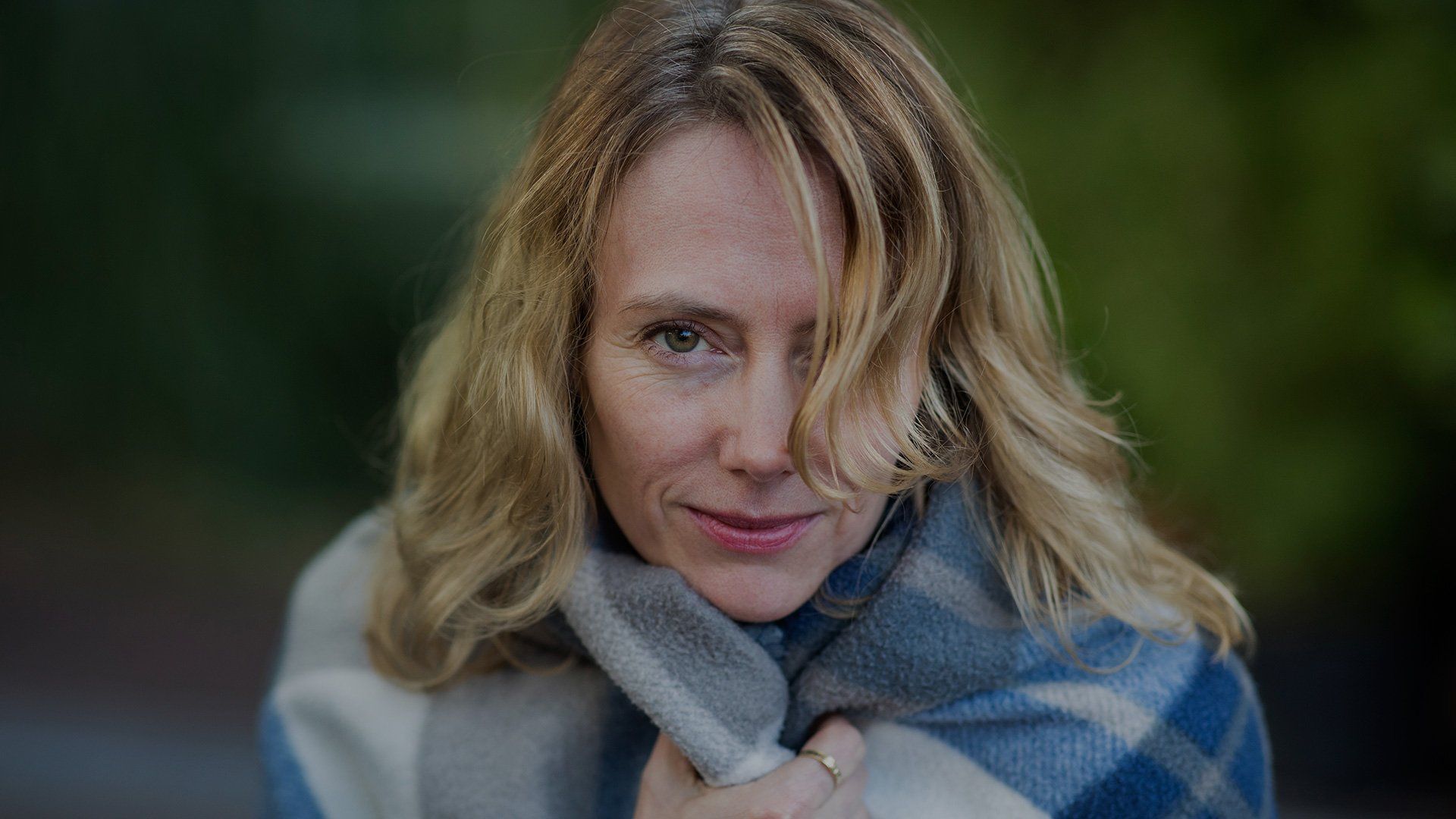 A portrait of a blonde woman outdoors, holding a soft blue checked fleece scarf close to her, taken with a Canon EF 85mm f/1.4L IS USM lens.